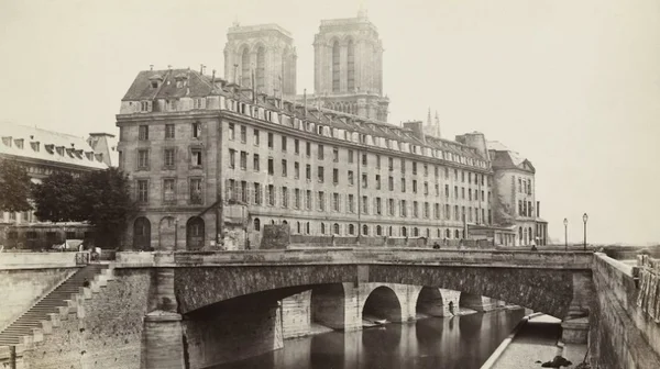 Photographie historique sépia montrant les tours jumelles de Notre-Dame de Paris derrière les bâtiments en bord de Seine, avec un pont de pierre en arche au premier plan près de l'Hôtel-Dieu
