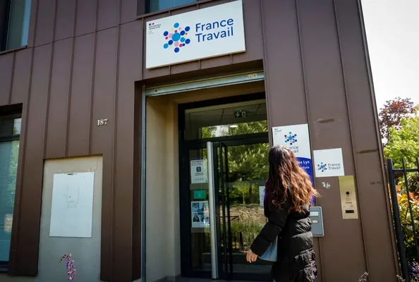 Femme aux longs cheveux bruns vue de dos entrant dans une agence France Travail au 187, bâtiment moderne aux portes vitrées avec signalétique visible entouré d'arbres