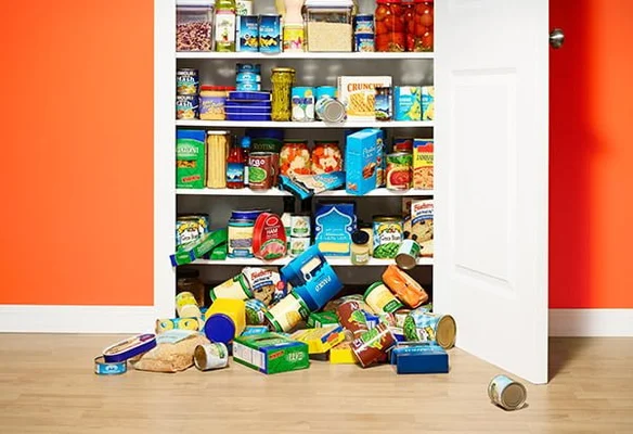 Placard garde-manger ouvert rempli de conserves, bocaux et boîtes sur les étagères, avec des provisions éparpillées sur le parquet devant la porte blanche