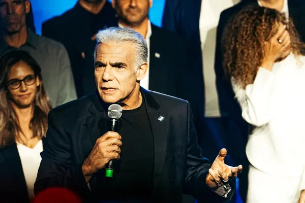 Yair Lapid, homme aux cheveux gris en veste sombre et chemise noire, parlant dans un micro lors d'un événement public entouré de plusieurs personnes