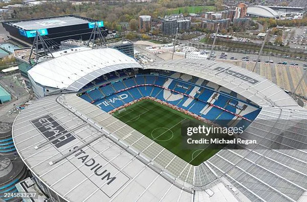 Vue aérienne du stade Etihad à Manchester avec son toit blanc incurvé, ses tribunes bleues aux inscriptions CITY et ETIHAD, la pelouse verte et le complexe environnant