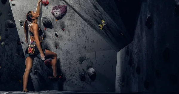 Grimpeuse avec une longue queue de cheval et un baudrier escaladant un mur artificiel gris parsemé de prises multicolores, attrapant une prise rouge en compétition de bloc en France