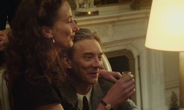 Un homme en costume tenant un verre et souriant, une femme penchée sur son épaule, dans un intérieur à la lumière chaleureuse, scène du film « Notre Salut » d'Emmanuel Marre