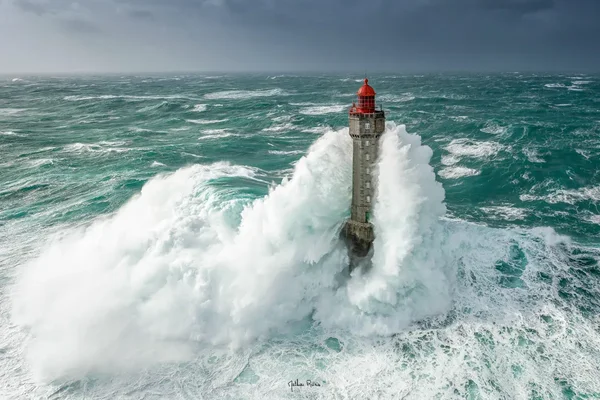 Le phare de la Jument à Ouessant en pleine tempête, une vague blanche massive s'écrasant contre le phare sous un ciel gris orageux
