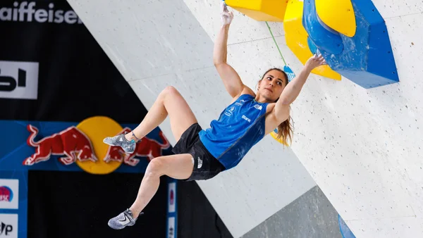 Grimpeuse en compétition de bloc féminin en intérieur, en pleine ascension sur un parcours bleu et jaune, avec les logos Red Bull et Raiffeisen en arrière-plan lors de la Coupe du monde