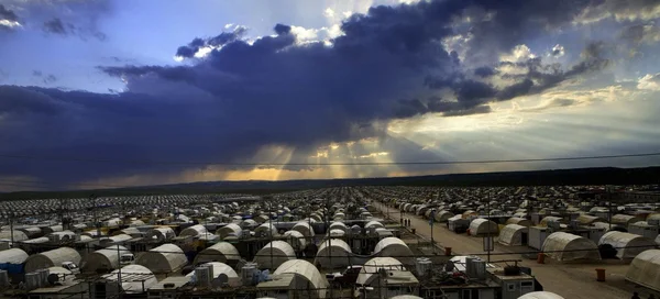 Vue panoramique du camp de réfugiés de Kabarto au Kurdistan irakien, avec des milliers de tentes blanches s'étendant jusqu'à l'horizon sous un ciel dramatique où les rayons du soleil percent les nuages sombres