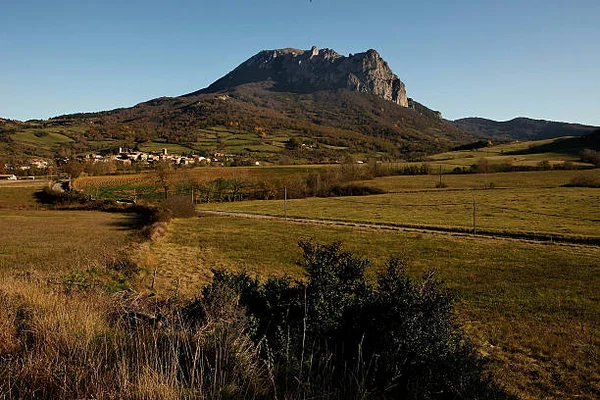 Vue générale du village de Bugarach dominé par son pic rocheux, avec maisons et champs vallonnés au premier plan sous un ciel bleu dégagé