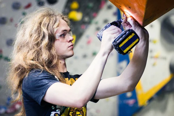 Personne aux cheveux longs ondulés portant des lunettes de sécurité et un t-shirt sombre utilisant une perceuse sans fil sur une structure en bois dans une salle d'escalade