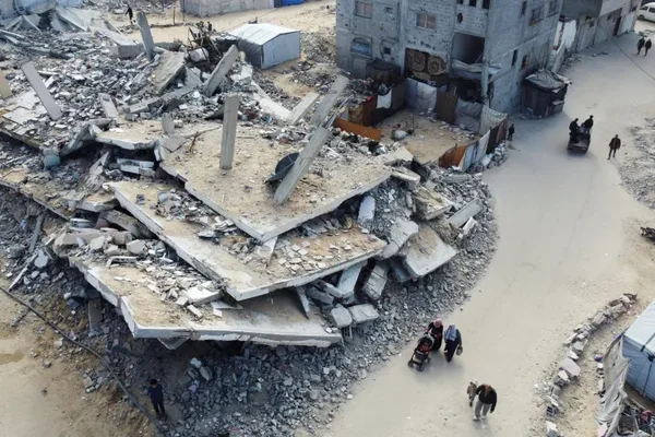 Vue aérienne d'un immeuble à plusieurs étages totalement détruit à Gaza, avec dalles de béton effondrées et gravats, le long d'une rue poussiéreuse où des Palestiniens et un âne marchent devant des bâtiments endommagés