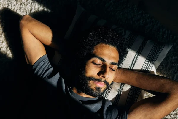 Homme barbu aux cheveux bouclés foncés allongé sur le dos les yeux fermés, mains derrière la tête, éclairé par une lumière chaude latérale sur fond sombre
