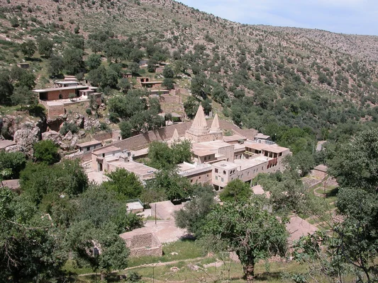 Vue en plongée d'un village yézidi en pierre niché parmi les arbres sur une colline rocheuse en Irak, centré sur un complexe de temples aux deux flèches coniques emblématiques