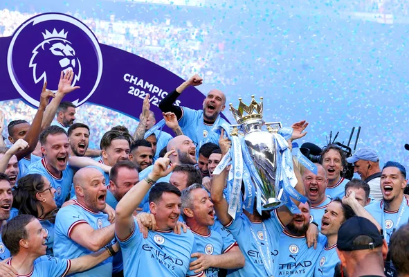 Les joueurs et le staff de Manchester City soulevant le trophée de Premier League 2022/23 sous une pluie de confettis bleus devant une bannière Champions
