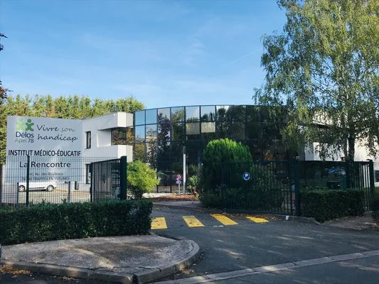 Entrée extérieure de l'Institut Médico-Éducatif La Rencontre de l'APEI Delos 78, avec panneau et logo, bâtiment moderne aux façades vitrées, portail et allée avec ralentisseurs jaunes