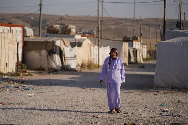 Un homme âgé yézidi déplacé irakien en robe violette claire et couvre-chef marchant sur un chemin de gravier dans le camp de Sharya pour personnes déplacées, avec tentes de fortune, poteaux électriques et collines en arrière-plan