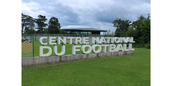 Entrée du Centre National du Football à Clairefontaine, surnommé « le Temple » du football français, avec des lettres blanches en 3D sur une pelouse, entouré de bâtiments modernes et d'arbres dans le Parc naturel régional de la Haute-Vallée de Chevreuse.