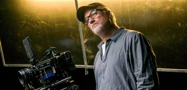 David Fincher portant casquette, lunettes et chemise sombre, debout à côté d'une caméra de cinéma professionnelle sur un plateau baigné d'une lumière dorée