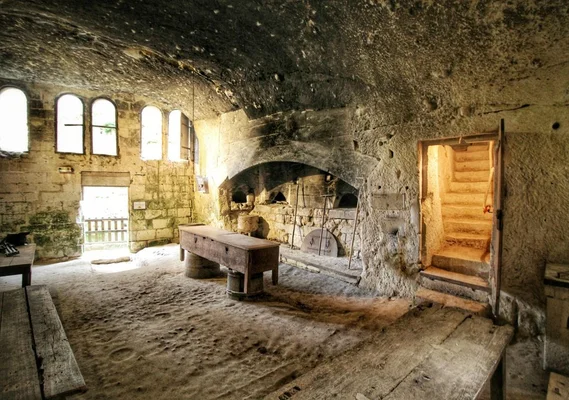 Intérieur en pierre du château de Brézé avec sol sablonneux, tables en bois, fenêtres en arc, fours maçonnés sous une voûte noircie et escalier en bois