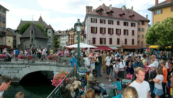 Foule compacte de touristes rassemblée autour du pont en pierre au bord du canal dans la vieille ville d'Annecy, avec bâtiments historiques et étal de vendeur dans une scène très animée