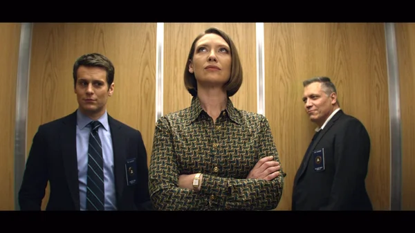 Les trois enquêteurs de la série Netflix Mindhunter sur les tueurs en série, une femme aux bras croisés entre deux agents en costume avec badges dans un ascenseur aux parois en bois