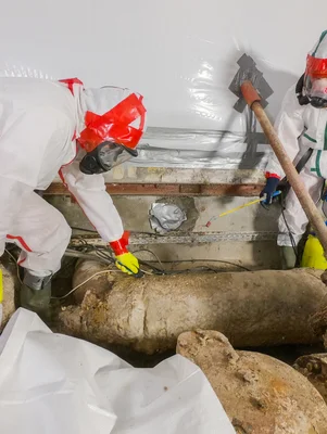 Deux travailleurs en combinaisons blanches intégrales avec masques respiratoires intervenant sur de grandes canalisations corrodées dans un espace souterrain confiné, risque cancérogène de l'amiante