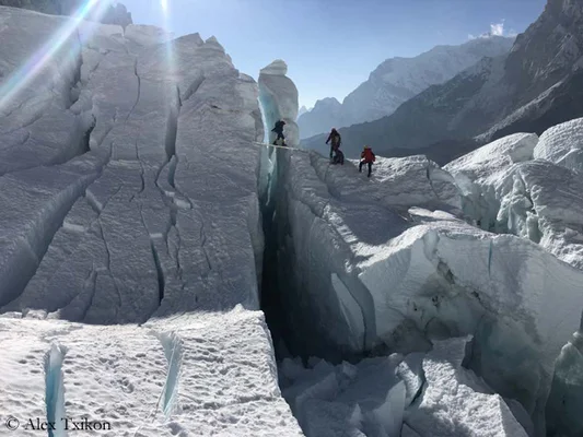 Trois alpinistes traversant une profonde crevasse glaciaire sur un pont de planches dans la cascade de glace du Khumbu sur le mont Everest, entourés de séracs fracturés et de sommets enneigés sous un soleil éclatant