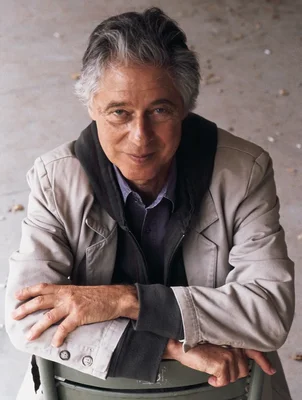 Portrait de l'écrivain américain Robert Littell, cheveux grisonnants, vêtu d'une veste claire sur un sweat à capuche sombre, penché en avant et les bras croisés sur le dossier d'une chaise.