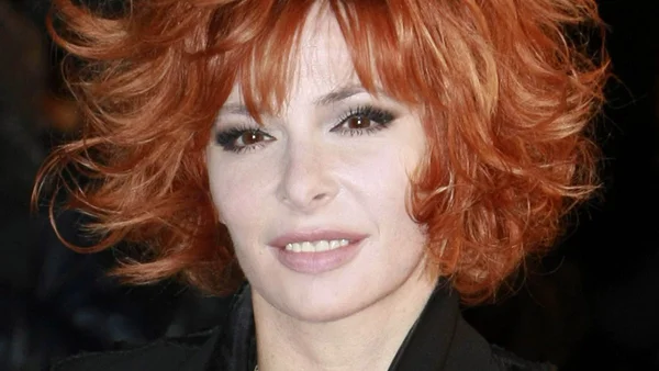 Portrait rapproché de Mylène Farmer avec sa chevelure rousse bouclée et volumineuse, maquillage des yeux foncé et tenue noire, esquissant un léger sourire face caméra