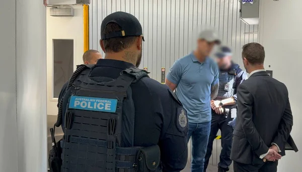 Ben Roberts-Smith, en chemise bleue et jeans, escorté par des officiers de la Police fédérale australienne dans un hôpital ou aéroport, avec un homme en costume derrière lui