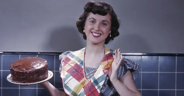Photographie vintage colorisée d'une femme au foyer des années 1950, cheveux courts bruns, robe à carreaux et tablier, souriante, tenant un gâteau au chocolat sur une assiette dans une cuisine.