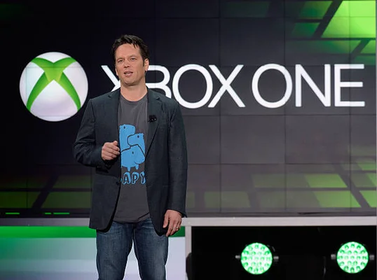 Phil Spencer, vice-président de Microsoft Game Studios, en costume et chemise, debout sur scène devant un écran affichant le logo Xbox One lors d'une conférence d'entreprise.