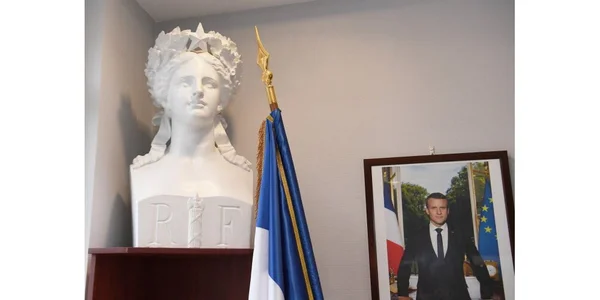Buste blanc de Marianne posé sur un support en bois, drapeau français et portrait encadré d'Emmanuel Macron accroché au mur dans une mairie.