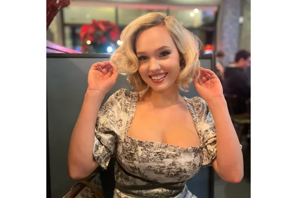 Estee Williams, influenceuse TikTok connue comme trad wife, cheveux blonds ondulés et robe à motifs, souriant les mains près des oreilles dans un intérieur lumineux.