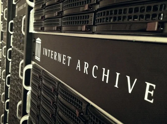 Photographie en gros plan d'une étagère de serveurs portant le texte blanc 'INTERNET ARCHIVE' et son logo, entourée de matériels informatiques empilés.