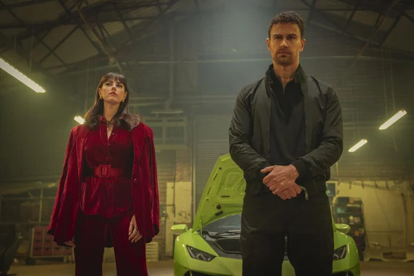 Kaya Scodelario et Theo James, acteurs principaux de la série The Gentlemen de Guy Ritchie sur Netflix, photographiés dans un garage devant une voiture de sport verte brillante avec le capot ouvert.