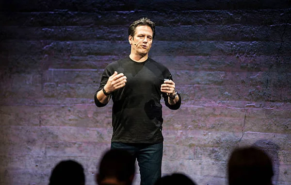 Phil Spencer, responsable de Microsoft Xbox et Microsoft Studios, en chemise noire et jean sombre, gesticule en parlant à un public sur scène, avec un mur de brique en arrière-plan.
