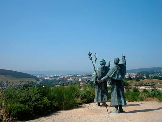 Deux statues en bronze de pèlerins en robes sur une colline, dominant une ville et un paysage sous un ciel bleu, évoquant le chemin de Compostelle.