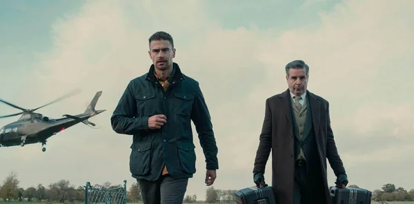 Deux hommes, l'un en costume et manteau formel tenant deux mallettes, l'autre en veste casual, marchent dans une prairie dégagée pendant qu'un hélicoptère atterrit en arrière-plan dans la série The Gentlemen de Guy Ritchie.