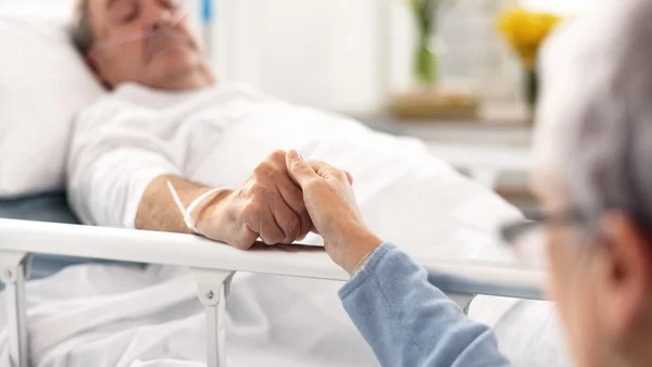 Gros plan sur deux mains jointes au-dessus de la barrière métallique d'un lit d'hôpital, une personne en fin de vie allongée dans le lit en arrière-plan