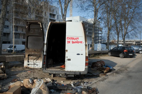 Camionnette blanche à portes arrière ouvertes garée sur un terrain près d'une route, avec des ordures et des graffitis rouges « ENFANT DE BOHÈME » sur la porte
