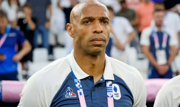 Thierry Henry, homme chauve avec une expression sérieuse, portant un polo blanc et bleu avec le logo de l’équipe olympique de France, assis dans un stade avec des spectateurs flous en arrière-plan.