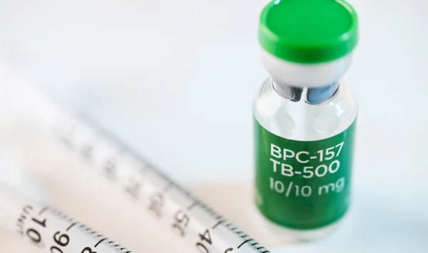 Gros plan sur un flacon en verre de peptides BPC-157/TB-500 de 10/10 mg, bouchon vert, posé sur une surface blanche à côté d'une seringue.