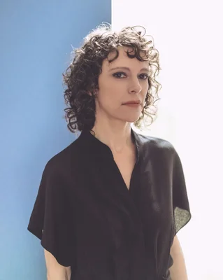 Portrait de la chanteuse Emily Loizeau, cheveux bruns bouclés et haut noir, devant un fond bleu et blanc.