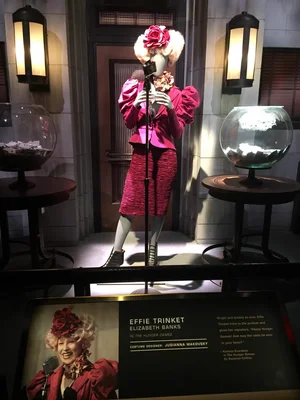 Mannequin costumé en Effie Trinket de Hunger Games, portant une tenue rose et un extravagant chapeau fleuri, exposé derrière un micro dans l'exposition au musée Rodin