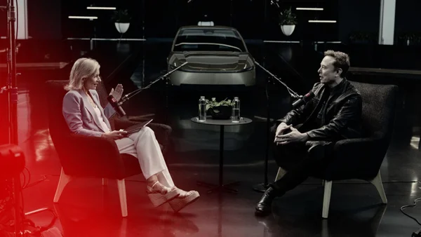 Elon Musk et son intervieweuse assis en face l'un de l'autre dans des fauteuils lors d'un entretien en studio, une voiture en arrière-plan
