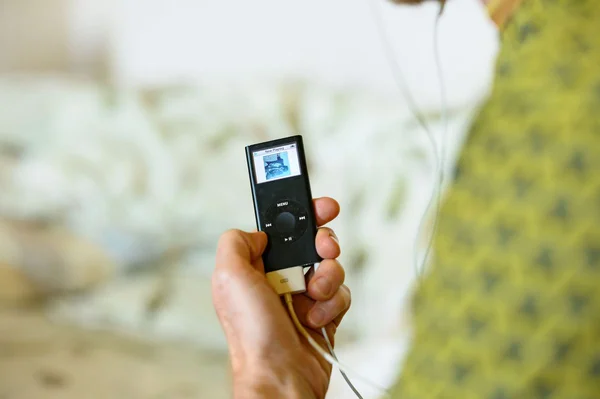 Main tenant un iPod Nano noir de 2006 de 8 Go, affichant une pochette d'album sur l'écran, relié à des écouteurs blancs