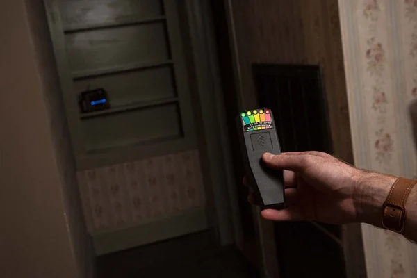 Main tenant un appareil de détection paranormale affichant un écran coloré dans un couloir faiblement éclairé, avec un petit dispositif électronique posé sur une étagère en arrière-plan