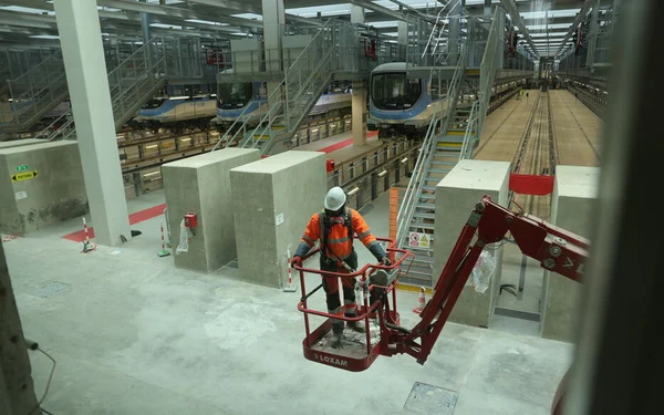 Ouvrier en gilet haute visibilité opérant une nacelle élévatrice rouge dans le dépôt de maintenance de la gare de Villiers-Champigny-Bry en construction, avec plusieurs trains garés sur les voies en arrière-plan