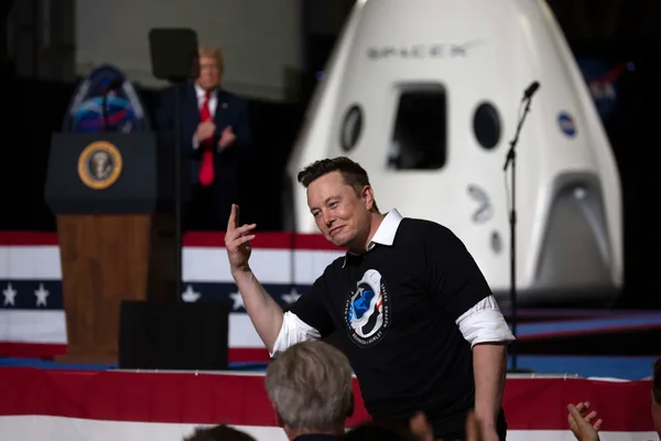 Elon Musk en t-shirt noir sur scène, gestiquant, devant une capsule spatiale SpaceX et un podium avec le sceau présidentiel américain