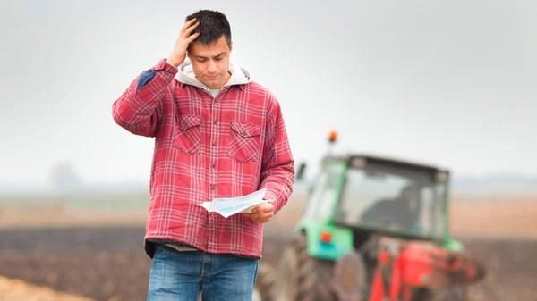 Agriculteur en chemise à carreaux rouges se tenant dans un champ, l'air inquiet tout en tenant un document, avec un tracteur vert en arrière-plan flou