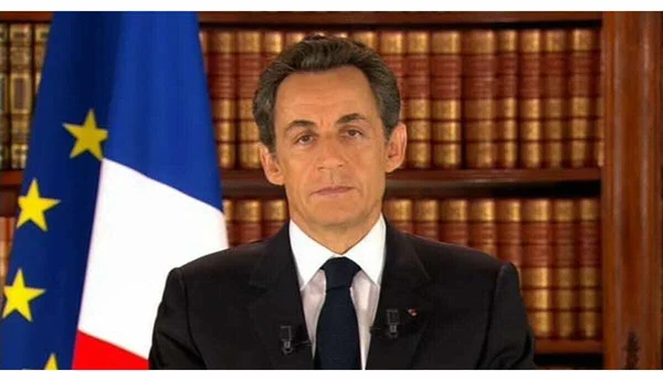 Nicolas Sarkozy en costume sombre et cravate s'adressant à la nation lors de son allocution télévisée, devant une bibliothèque avec le drapeau français à sa gauche